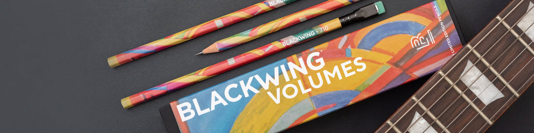 Three multicolored pencils and a blackwing volumes pencil box on a dark surface.