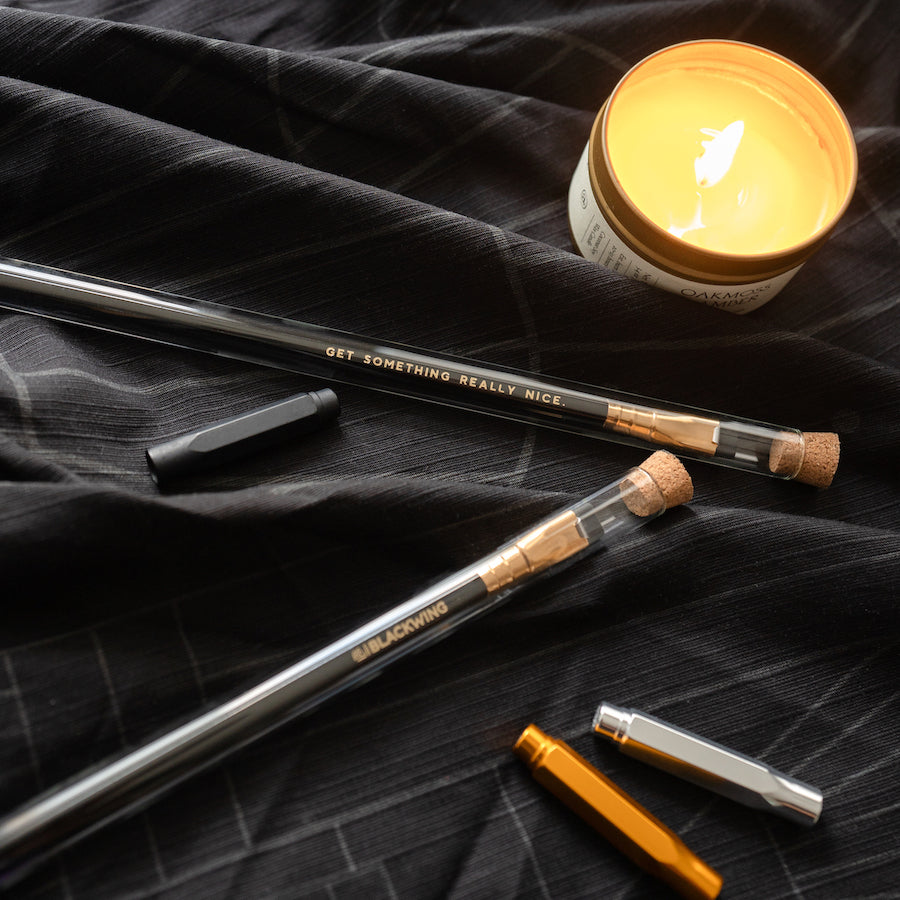 Two Blackwing Gift Card Pencils and a candle are displayed on a black cloth.