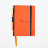 An orange Blackwing x Audubon birding journal with a black elastic closure band, a matching orange ribbon bookmark, and a pencil tucked into the side, photographed on a clean white background.