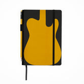 A black hardcover notebook with a large mustard yellow guitar-shaped graphic on the cover, shown closed with a black elastic closure. A yellow pencil is tucked into the side holster, and a black ribbon bookmark hangs from the bottom, on a clean white background.