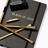 Black notebook with 'Class of 2026' gold foil stamped imprint with gold pencils, a black sharpener, and a gold point guard all on top of a white background
