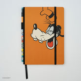 An orange hardcover notebook by Blackwing featuring a cartoon Goofy illustration on the front, with a black elastic closure band and a matching black ribbon bookmark. A colorblock pencil with Goofy graphics is tucked into the left side.
