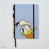 A light blue hardcover notebook featuring Donald Duck's face on the cover, with a black elastic closure band and a matching black ribbon bookmark. A matching colorblock pencil with colorful Donald Duck illustrations is tucked into the left side of the notebook.