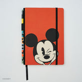 A red Mickey Mouse notebook with a black elastic closure band, featuring a winking Mickey face on the cover and a pencil holster with colorful matching pencil visible on the side, photographed on a light gray background.
