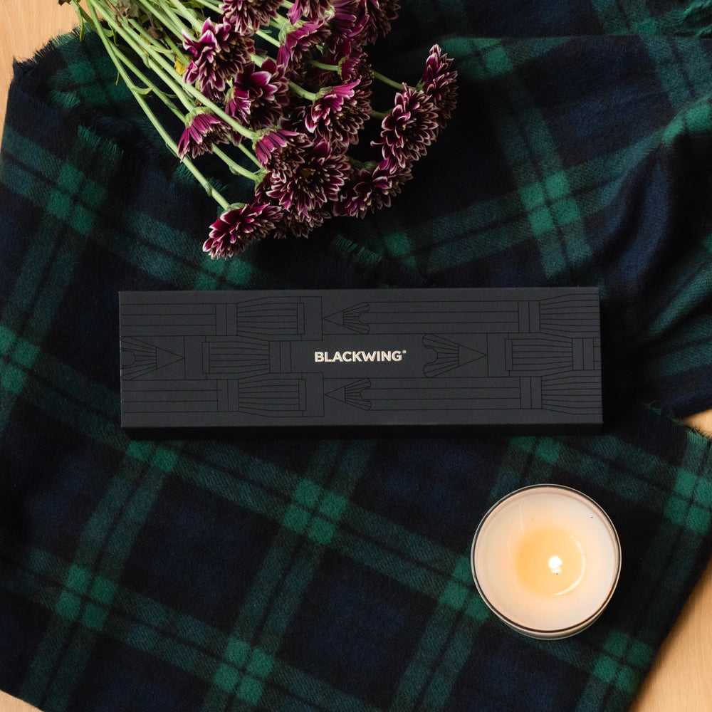 A Blackwing Pencil Essentials Set sits beside a plaid blanket, surrounded by flowers.