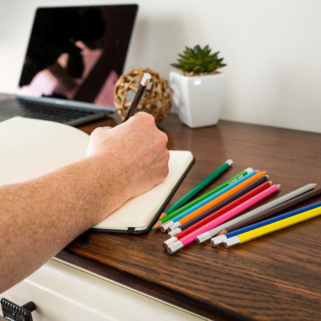 A person writing on a desk with Blackwing Colors (Set of 12) and a laptop, adding a creative touch to their work.