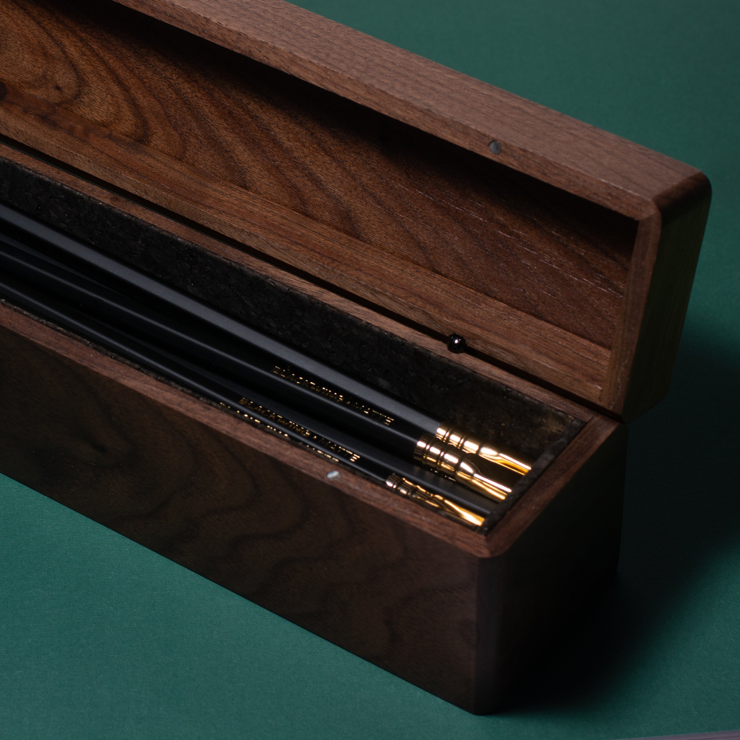 A Blackwing Walnut Box containing Blackwing pencils rests on the green surface.