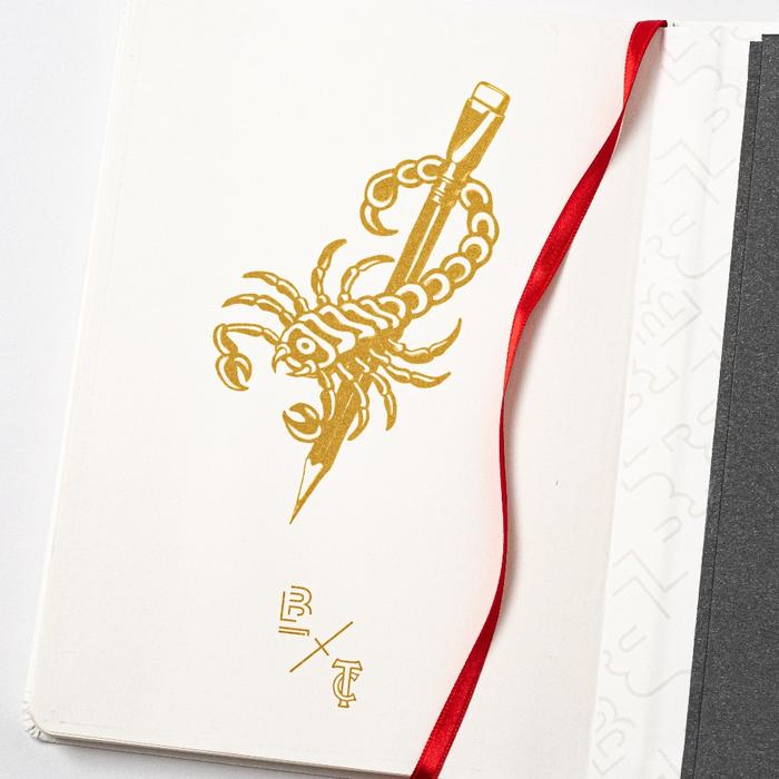 A Blackwing x Timeless Coffee Slate Notebook with a scorpion on it and a red ribbon.