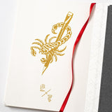 A notebook with a scorpion on it and a red ribbon, perfect for jotting down thoughts during a Blackwing x Timeless Coffee Bundle break.