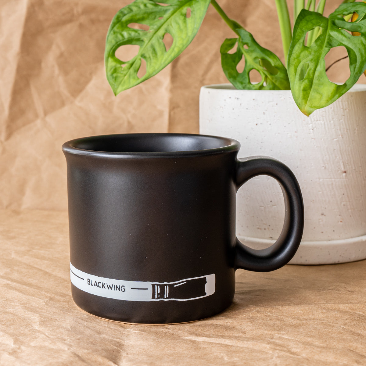 A black mug with a plant next to Blackwing Coffee Mug.