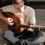 A man wearing a Blackwing "B" Blueprint T-Shirt plays an analog guitar while sitting on the floor.