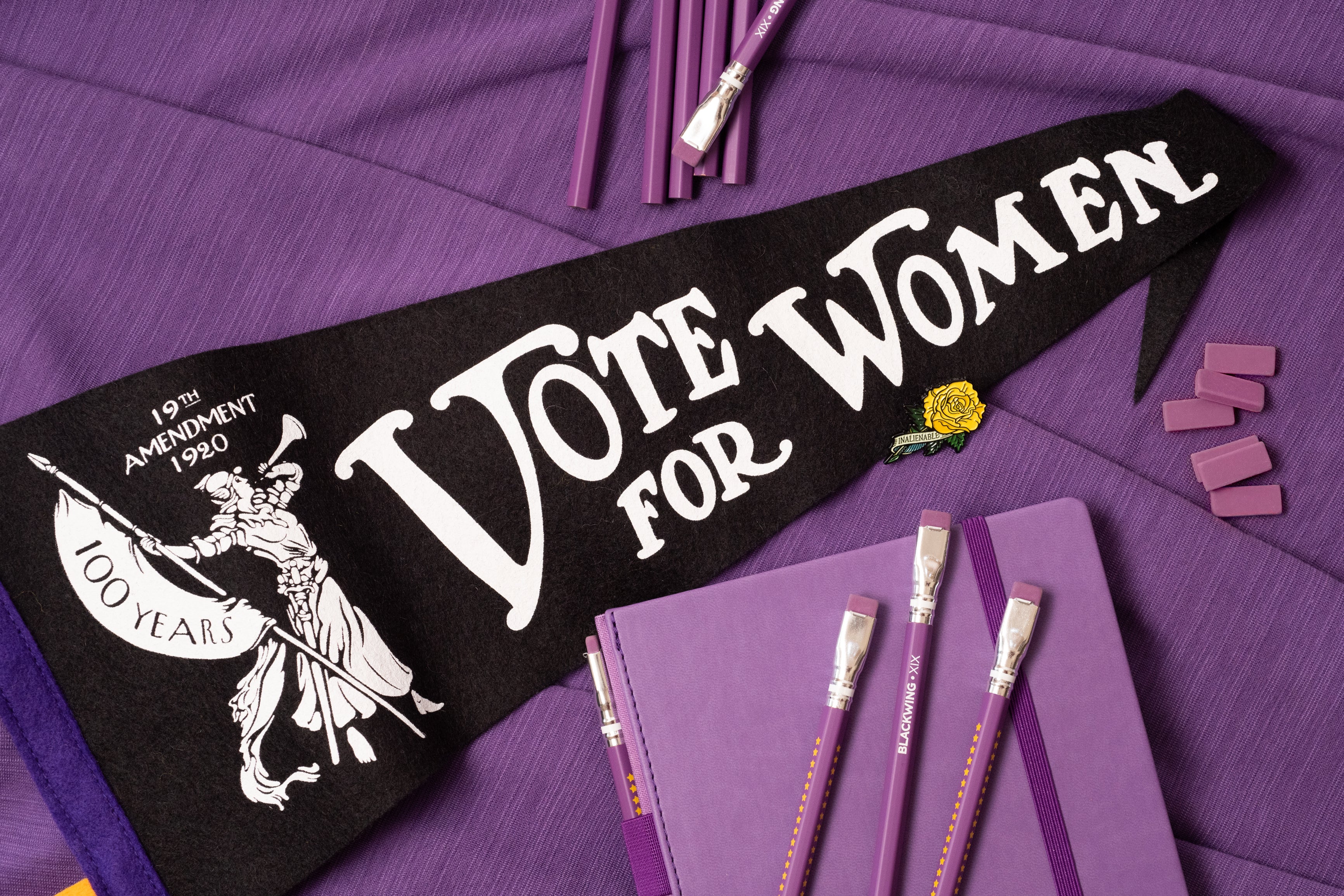 A limited edition Blackwing XIX “Vote for Women” Pennant alongside symbols of women's suffrage, a commemorative badge, and Blackwing XIX purple-themed stationery arranged on a purple background