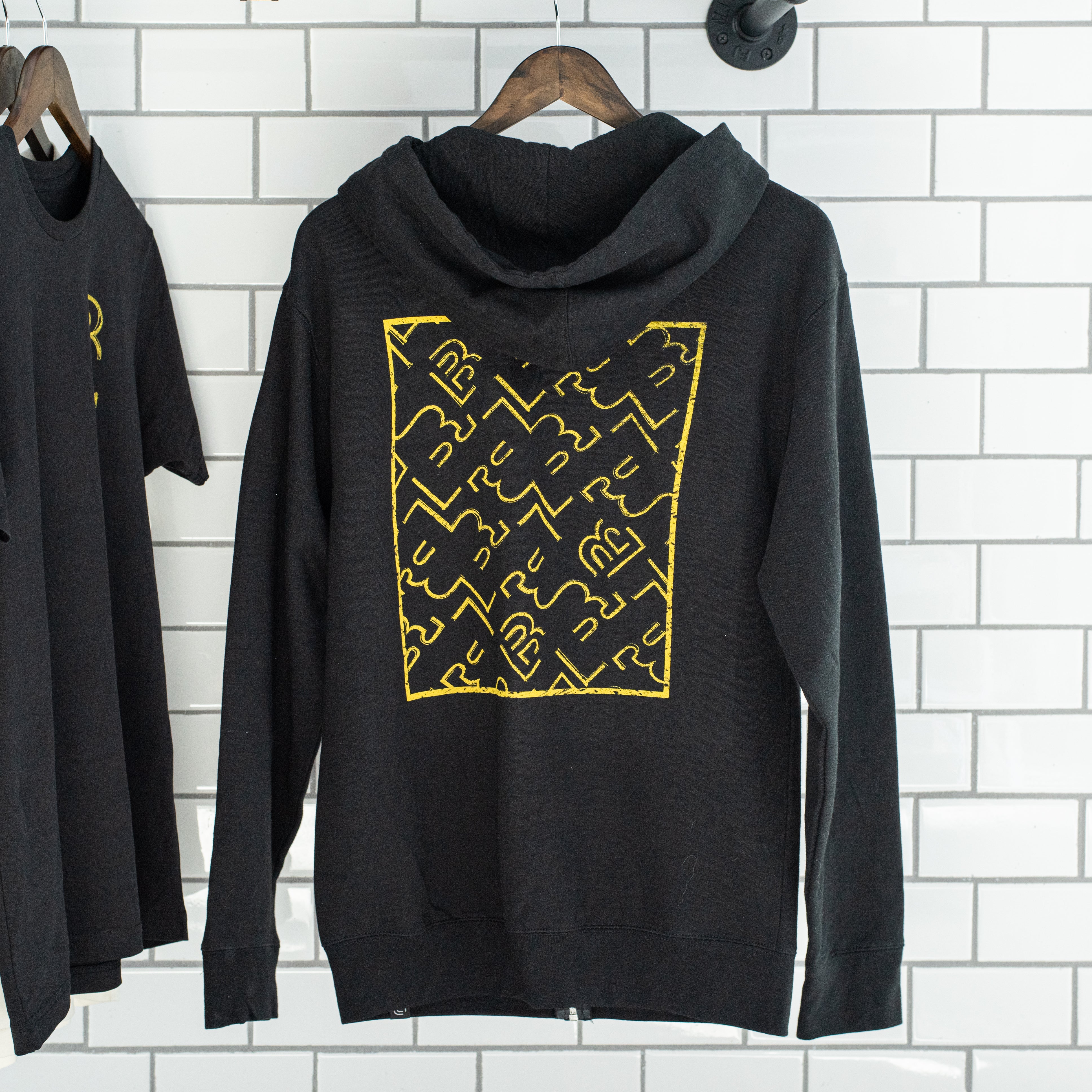 A Blackwing Sketch Zip-up Hooded Sweatshirt with a yellow design hangs on a brick wall from the Winter 2019 Blueprint capsule.
