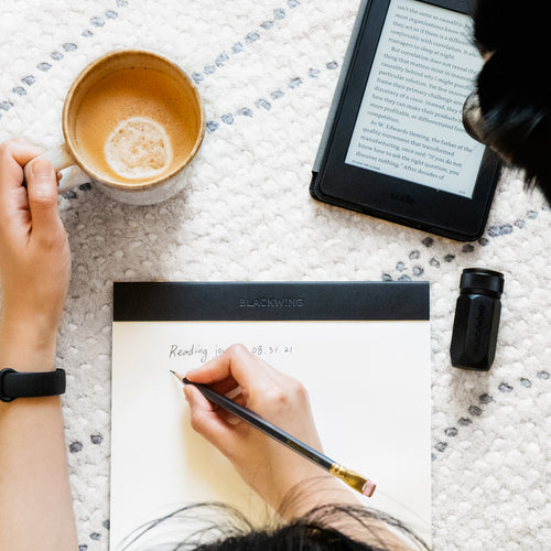 Person writing with Blackwing pencil on notepad with coffee