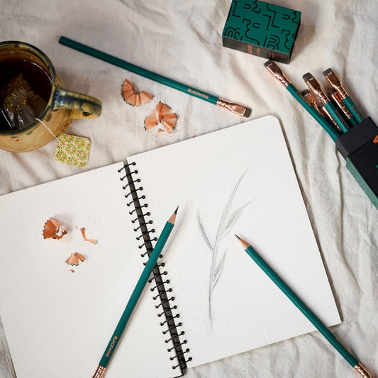 A Blackwing spiral notebook open with a drawing of a plant. Blackwing Matte Green Pencils surrounding the notebook and cup of tea on the table.