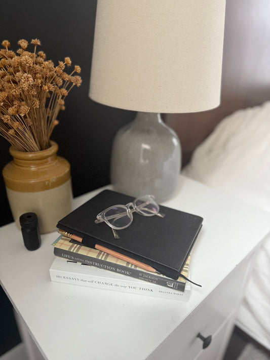 Aesthetic image of nightstand with a lamp, pottery with dried florals, books, eyeglasses, a blackwing journal, a blackwing pencil, and a blackwing sharpener