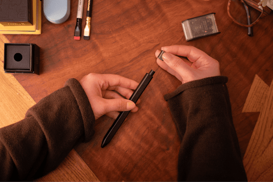 person's hands holding a matte black pen, removing the cap over a wood desk full of pencils, a sharpener, and other accessories
