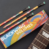 A collection of Blackwing Volume 710 (Set of 12) pencils alongside their packaging box with a striped color pattern, displayed on a dark surface next to a stringed musical instrument associated with Jerry Garcia.