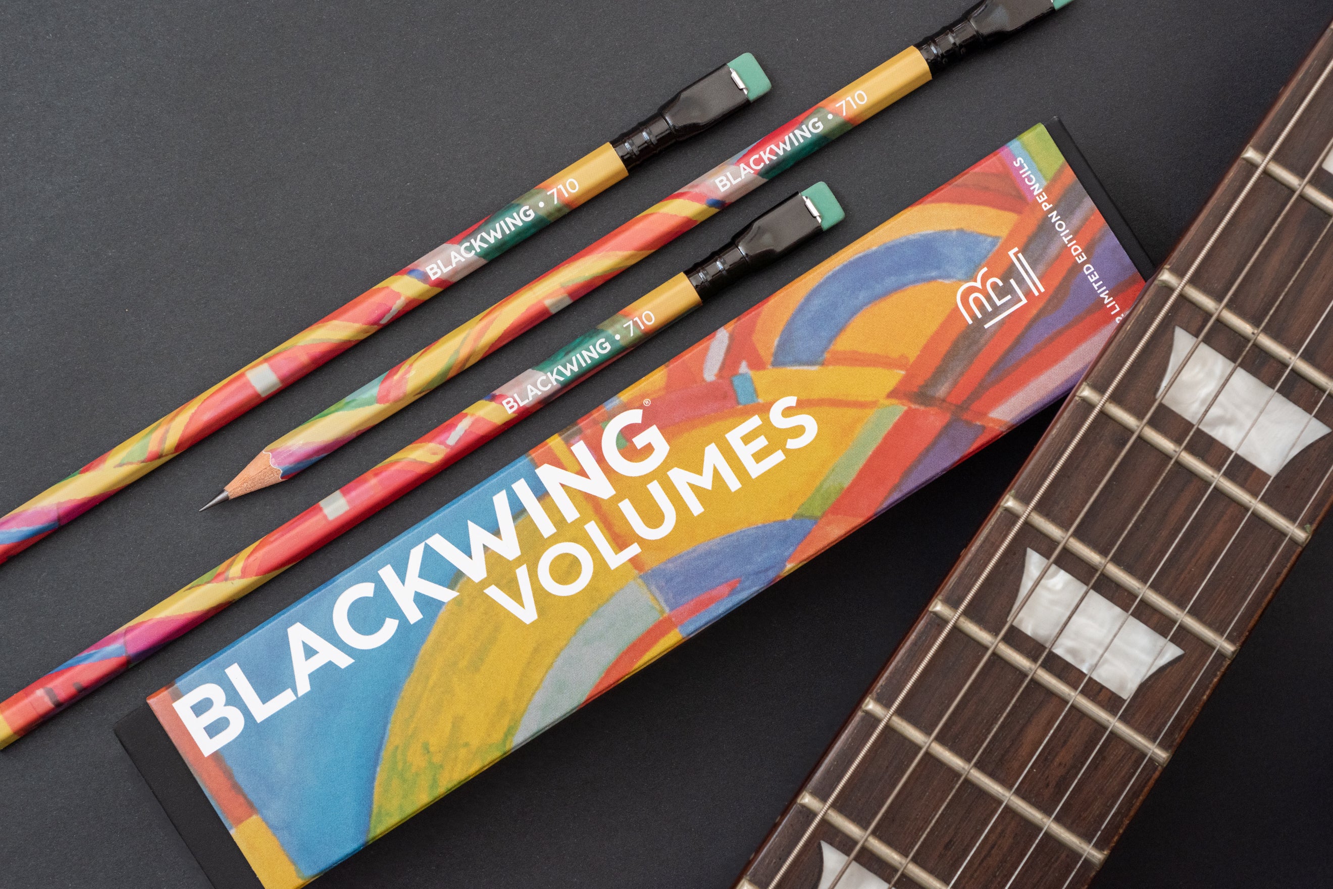 Three Blackwing Volume 710 (Set of 12) pencils and a pencil box with a colorful Grateful Dead design next to a guitar fretboard on a dark surface.