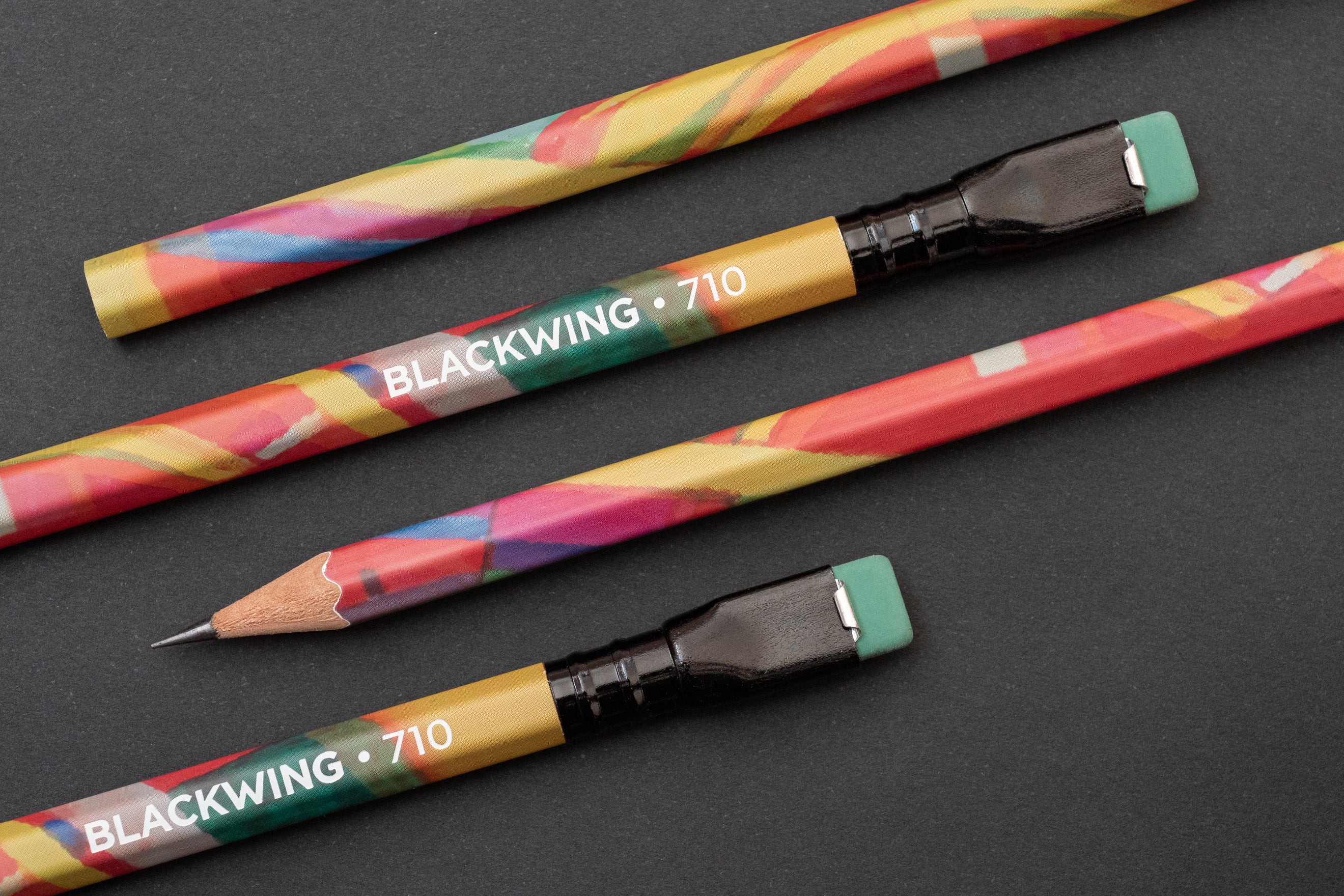 Multicolored Blackwing Volume 710 pencils with erasers, one sharpened, on a dark surface.