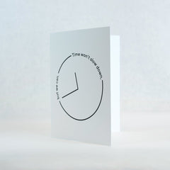 white greeting card featuring a black thin outlined clock with the words "time won't slow down, but we can" text along the clock outline.  