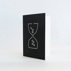 black greeting card featuring a white outlined hourglass and "slow down" text as the "sand". Inside of card is white.