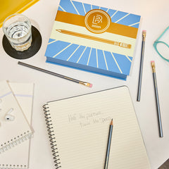 A styled flat-lay of stationery items on a white surface, including a blue and gold branded box with 'B' Blackwing emblem on the front. Around it is pencils, an open lined notebook with handwritten text 'half the pressure, twice the speed', a glass of water, earbuds, and eyeglasses.