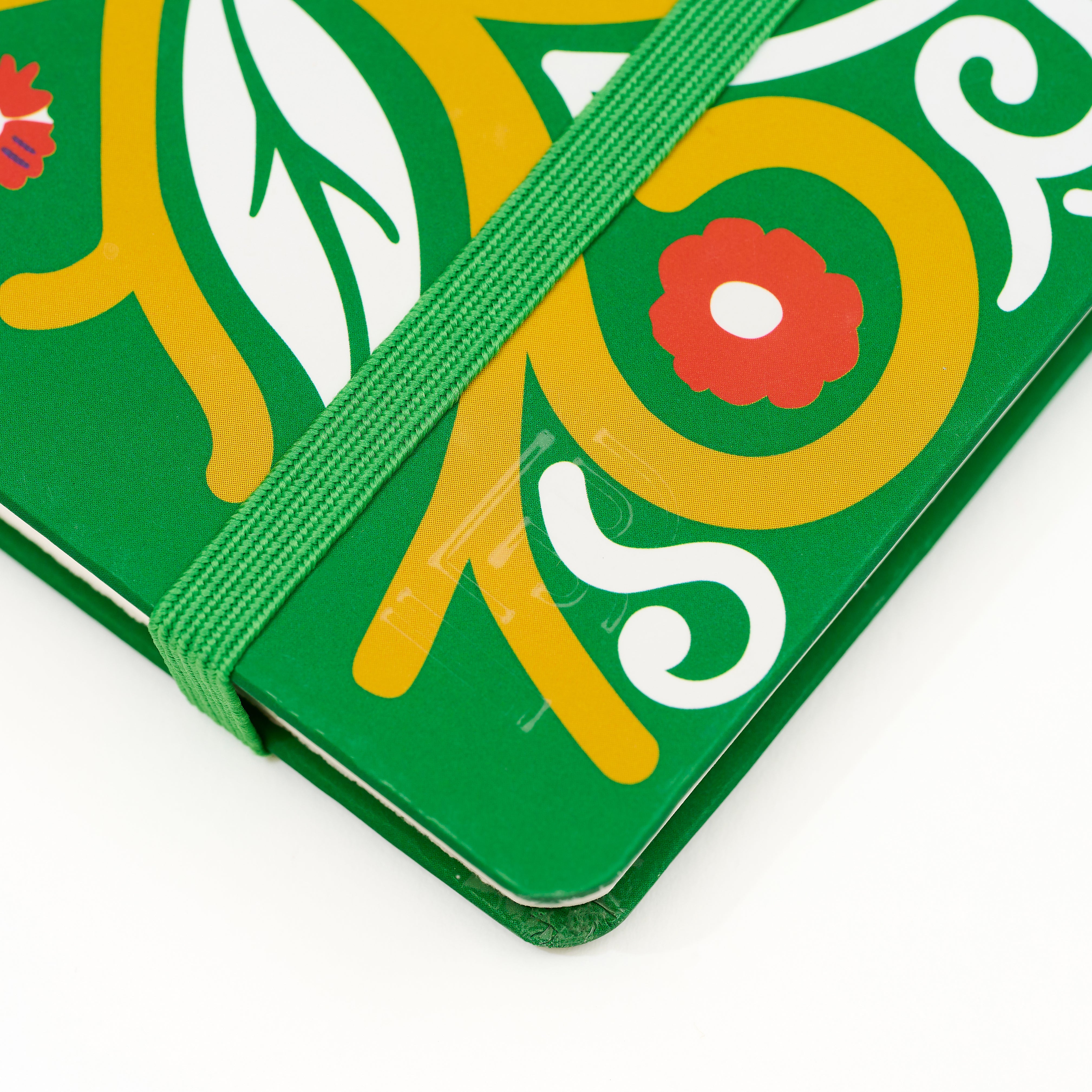 A close-up of a green hardcover notebook or journal with a colorful printed floral and leaf pattern in yellow, white, and red, secured with a matching green elastic closure band. The corner features the Blackwing 'B' emblem stamped into the notebook.