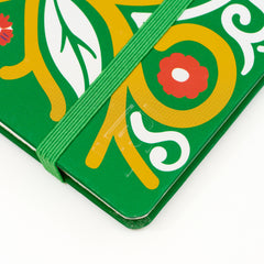 A close-up of a green hardcover notebook or journal with a colorful printed floral and leaf pattern in yellow, white, and red, secured with a matching green elastic closure band. The corner features the Blackwing 'B' emblem stamped into the notebook.