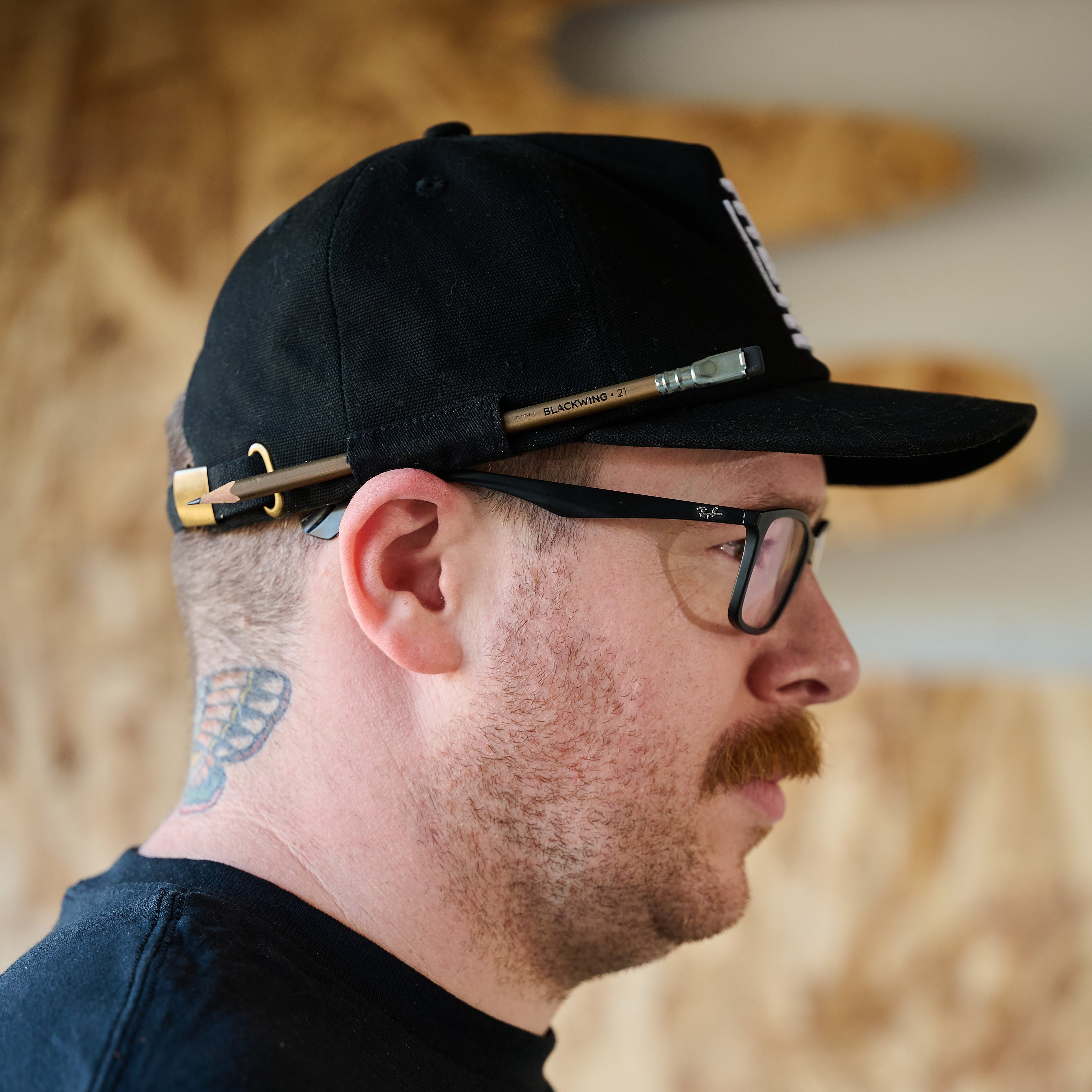 Man with glasses wearing a black hat with white Blackwing emblem embroidered on the front. The cap features a loop on the side to hold a pencil, featuring Blackwing Volume 21.