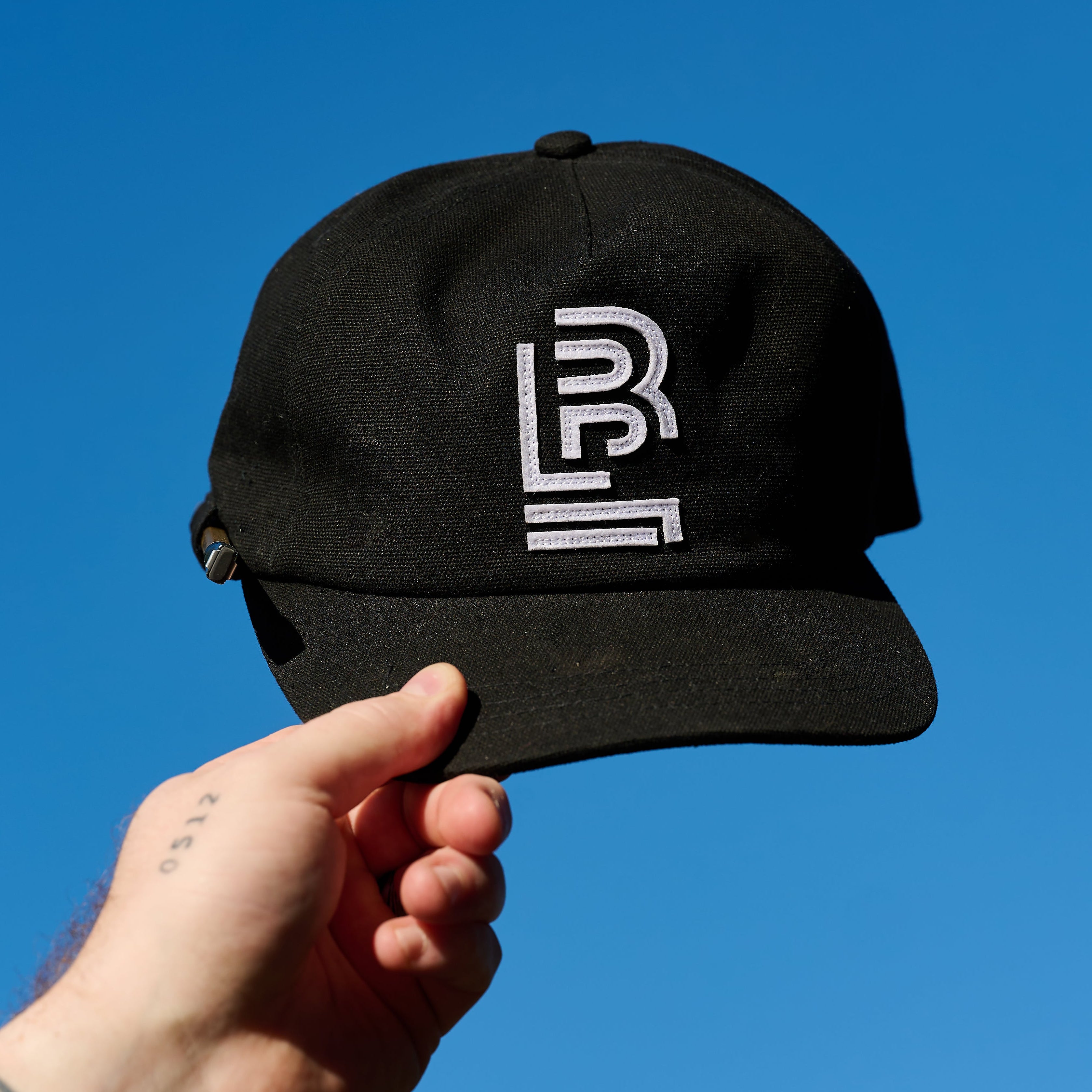 Person's hand holding a Black cap up in the sky. The cap features a white embroidered Blackwing emblem on the front and the side of the cap can hold your favorite pencil.