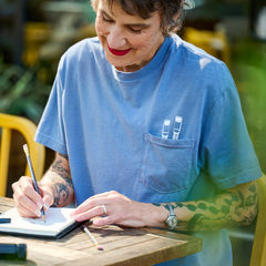 Woman wearing a blue t-shirt sitting at a table writing in a spiral notebook with a pencil.