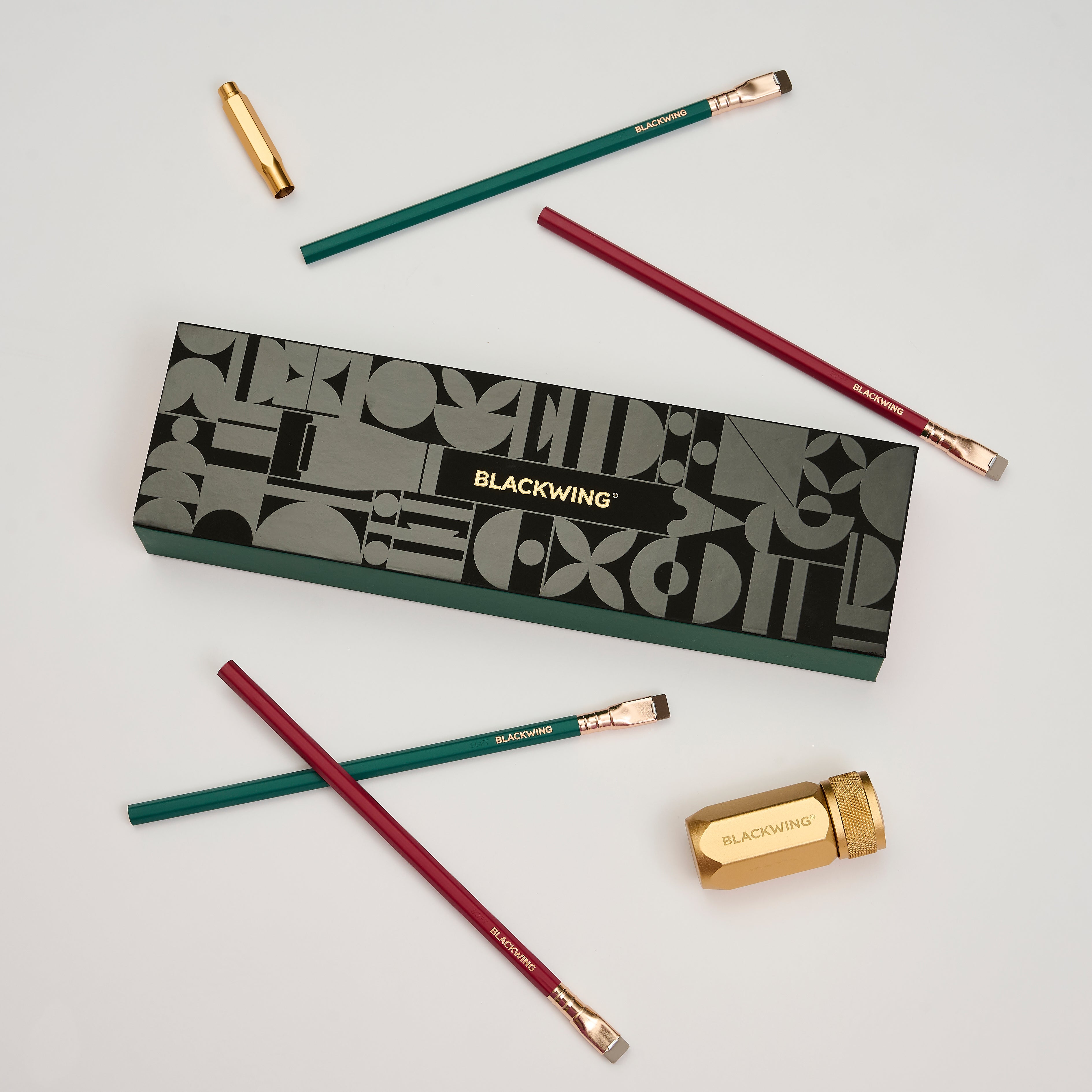 Blackwing Holiday Essentials Gift Set Box surrounded by the contents, including 2 green pencils, 2 red pencils, a gold sharpener, and a gold point guard on a white background.