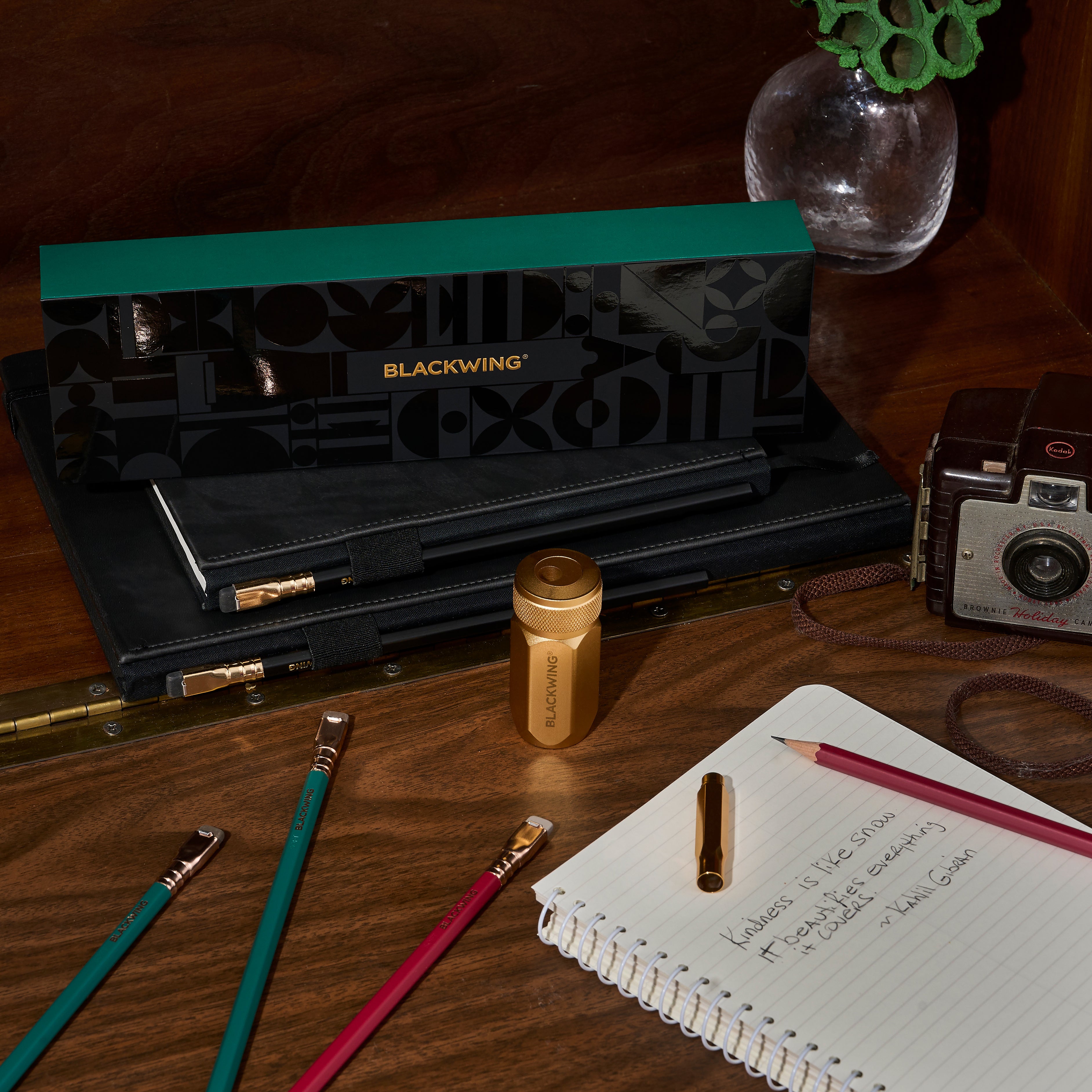 Blackwing Holiday Essentials Gift Set Box surrounded by the contents, including 2 green pencils, 2 red pencils, a gold sharpener, and a gold point guard on a wood desk with a spiral notebook and old fashion camera.
