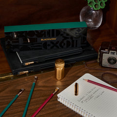 Blackwing Holiday Essentials Gift Set Box surrounded by the contents, including 2 green pencils, 2 red pencils, a gold sharpener, and a gold point guard on a wood desk with a spiral notebook and old fashion camera.