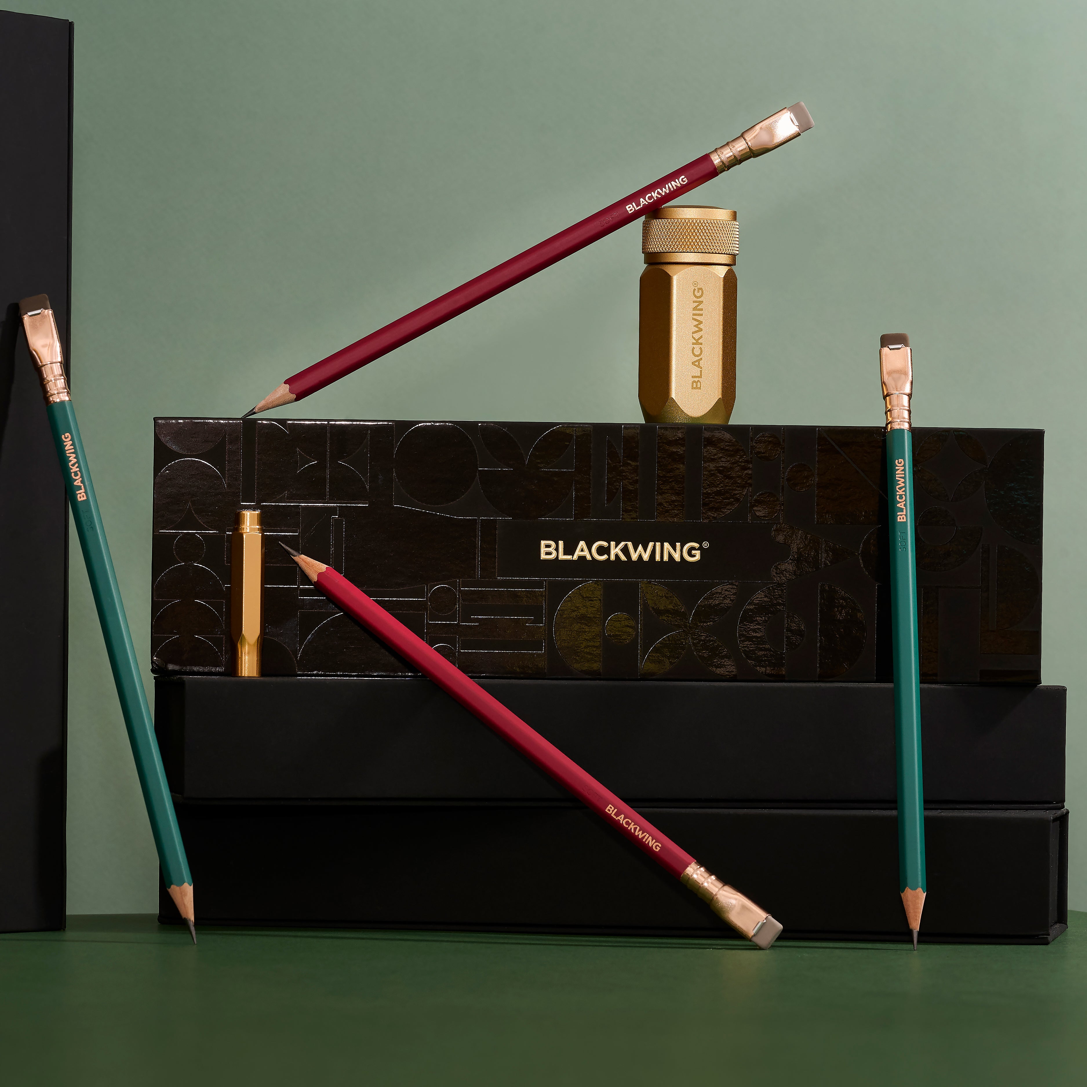 Blackwing Holiday Gift Set Box surrounded by the contents, including 2 green pencils, 2 red pencils, a gold sharpener, and a gold point guard on a green background.