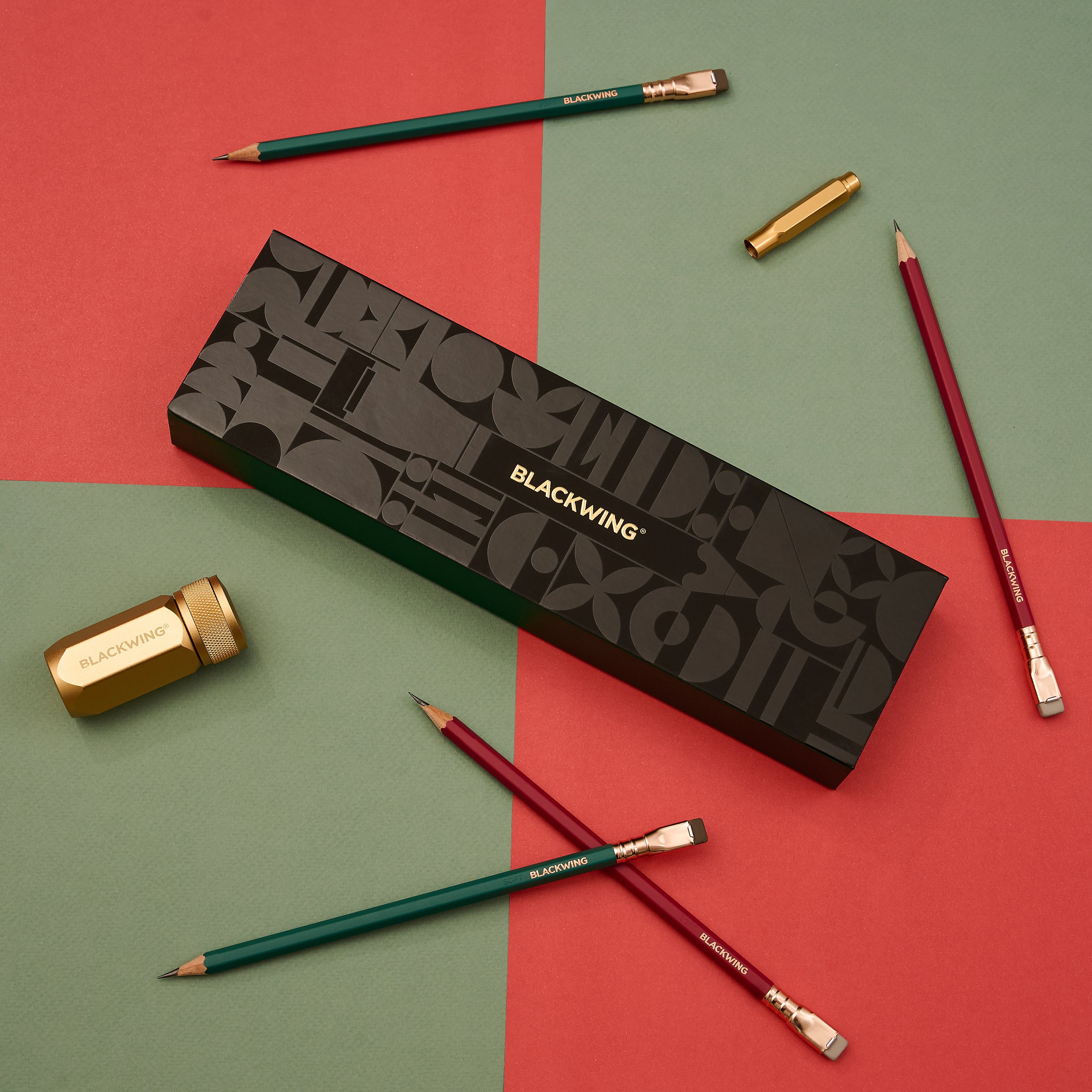 Blackwing Holiday Essentials Gift Set Box surrounded by the contents, including 2 green pencils, 2 red pencils, a gold sharpener, and a gold point guard on a red and green checkered background.