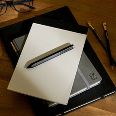 A blackwing rollerball pen lays on top of a dot-grid notepad and two other hardcover notebooks on top of a wooden desk, which also features two blackwing pencils and black glasses.