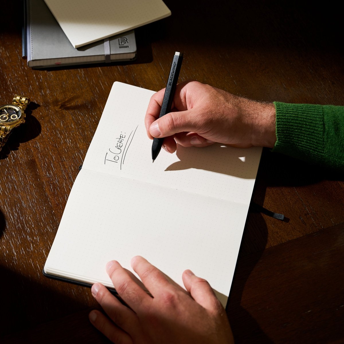 hand holding a pen in right hand that has written "To Create" in black ink into a notebook on a wood desk with