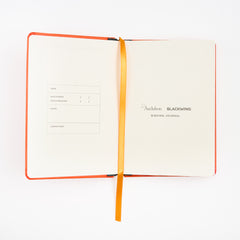 An open Audubon x Blackwing birding journal with cream-colored pages, an orange cover, and a gold ribbon bookmark, displayed on a clean white background.