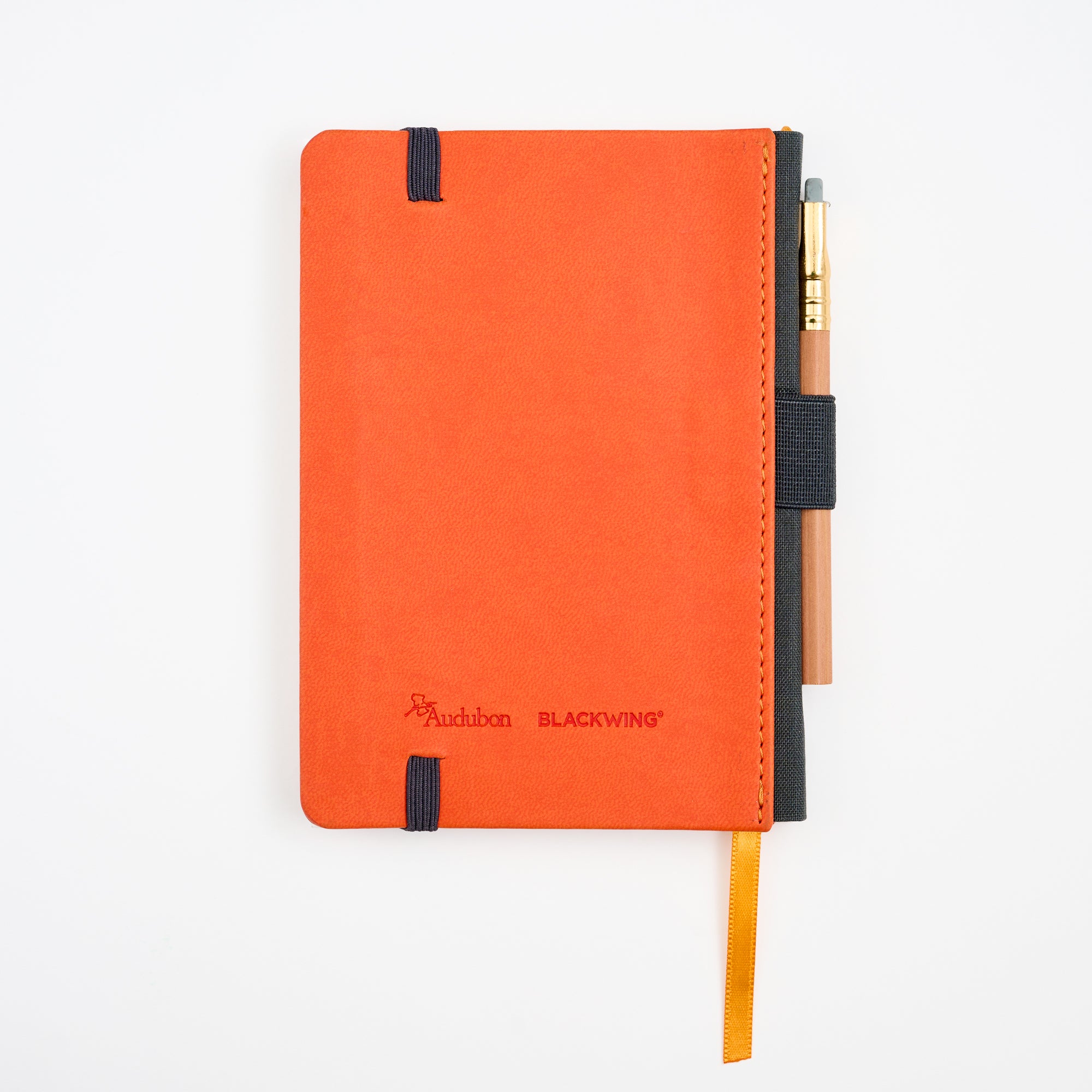 An Audubon x Blackwing hardcover notebook in vibrant orange, featuring a black elastic closure, a gold-orange ribbon bookmark, and a short pencil tucked into the black side holster, all photographed on a clean white background.
