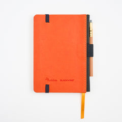 An Audubon x Blackwing hardcover notebook in vibrant orange, featuring a black elastic closure, a gold-orange ribbon bookmark, and a short pencil tucked into the black side holster, all photographed on a clean white background.