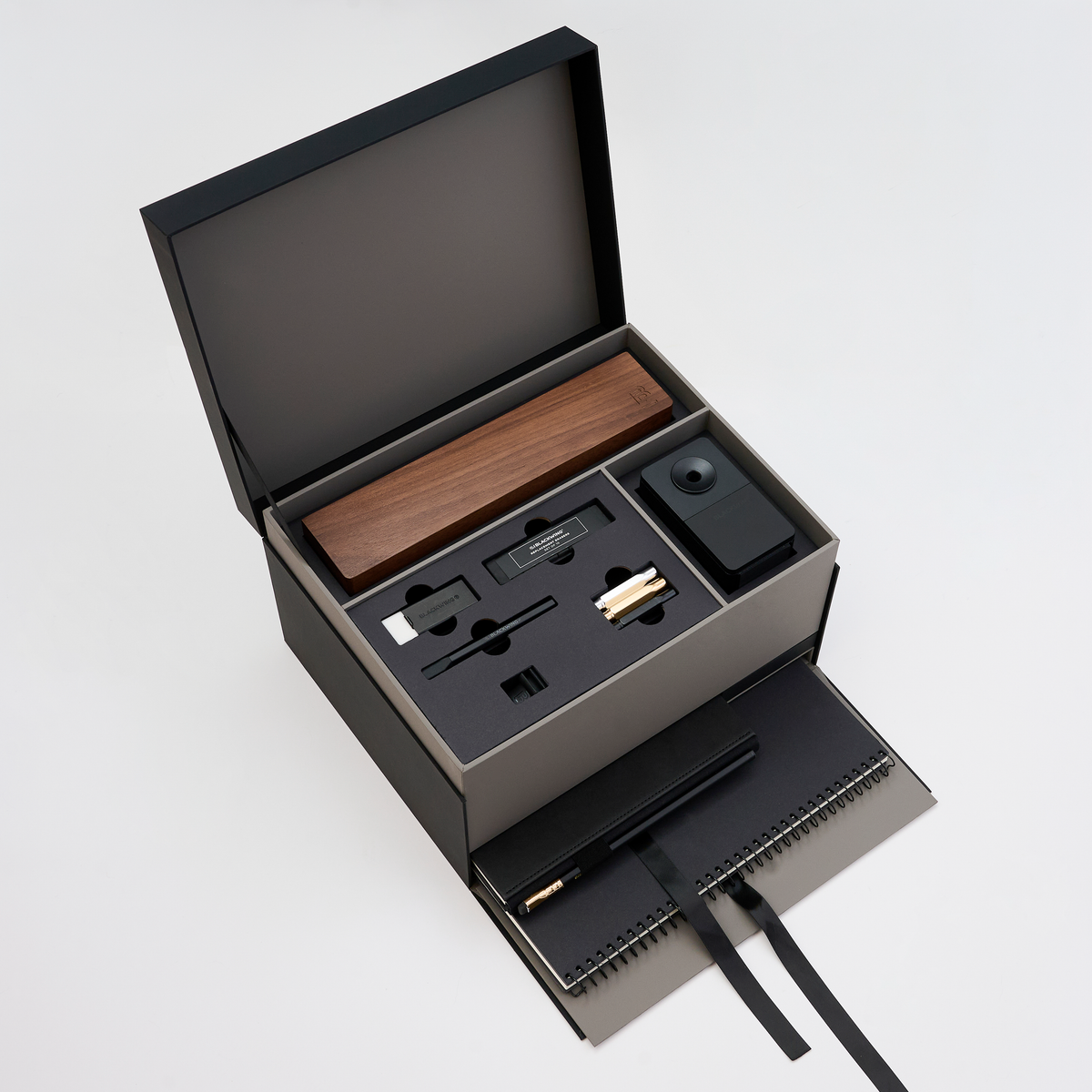 An open black gift box on a light gray background, revealing a wooden pencil box display set, blackwing handheld eraser, blackwing replacement erasers, blackwing point guards, and blackwing notebook clip on the top layer, and a large black spiral bound notebook and medium hardcover notebook with a black pencil in the holster in a lower pull-out drawer with a ribbon string.