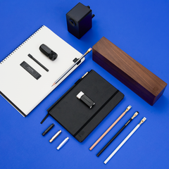 A flat-lay arrangement of blackwing stationery and office accessories on a bright blue background, including a white paper spiral notebook, a black hardcover notebook, several pencils, a black pencil sharpener, and a long rectangular wooden box.