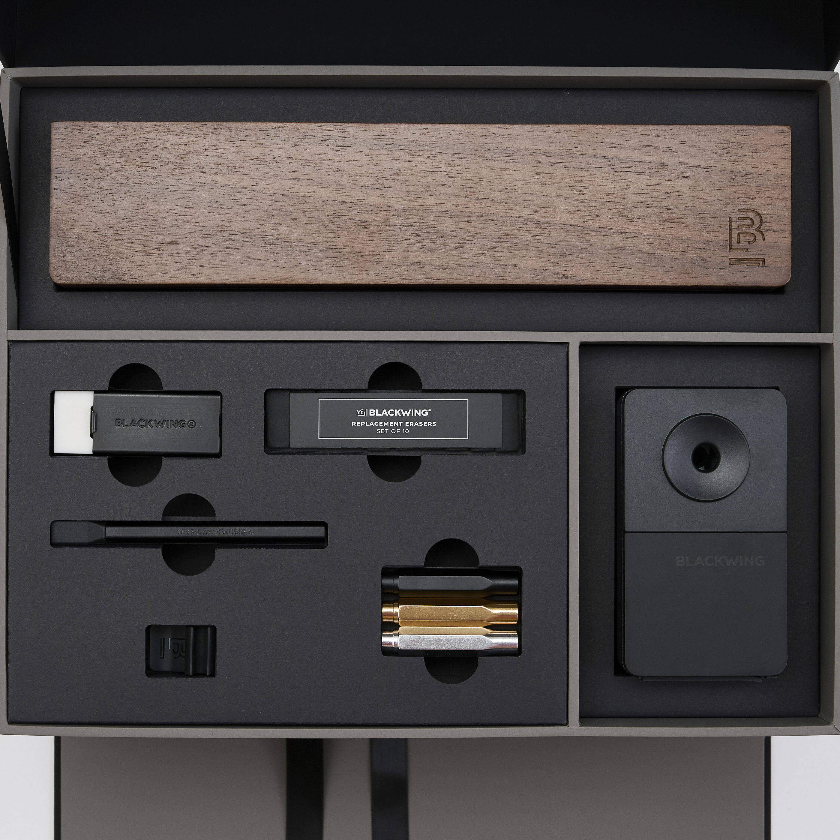 Blackwing stationery items set in a premium presentation box, featuring a wood display case, matte black pencil extender, black and white handheld eraser, black replacement erasers, a black desktop sharpener, a black notebook clip, and silver, gold, and black point guard pencil protectors.