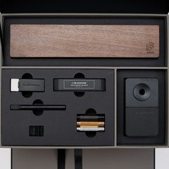 Blackwing stationery items set in a premium presentation box, featuring a wood display case, matte black pencil extender, black and white handheld eraser, black replacement erasers, a black desktop sharpener, a black notebook clip, and silver, gold, and black point guard pencil protectors.