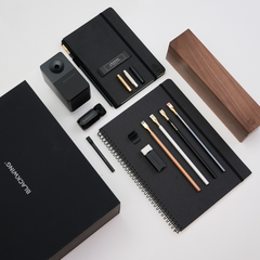 A black stationery and office accessory set arranged on a light gray background, including notebooks, a wood pencil display case, a pencil sharpener, a pencil extender, and other accessories and branded packaging. The visible brand text reads BLACKWING.