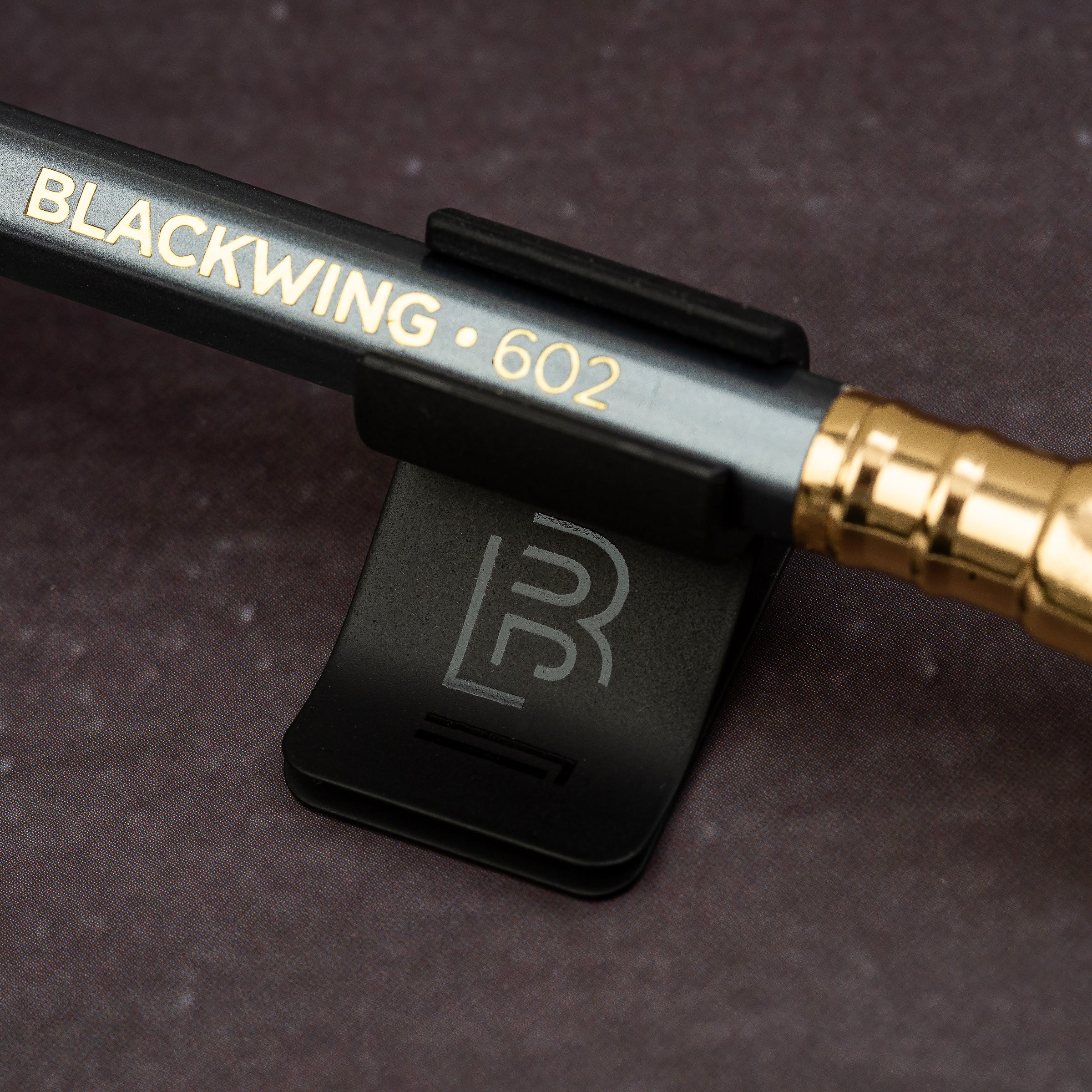 Blackwing Notebook Clip in black with a "B" emblem holding a Blackwing 602 pencil