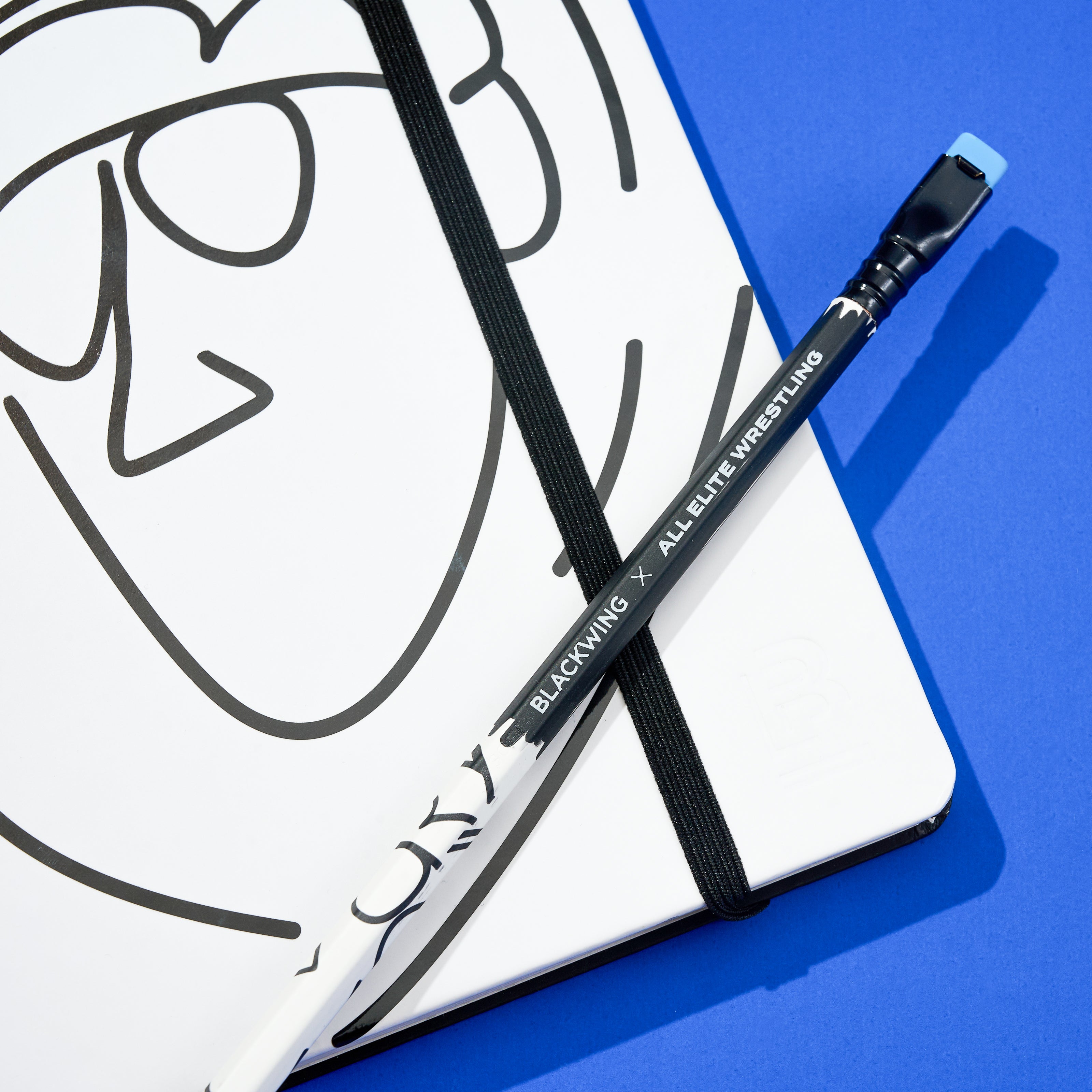 Blackwing x AEW "Freshly Squeezed" Orange Cassidy Notebook with a minimalist black line drawing of a face on a white cover with a matching pencil on top, all on a blue background