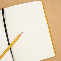 An open blank notebook with a yellow cover and black ribbon bookmark, placed on a tan background with a yellow pencil resting diagonally across it.